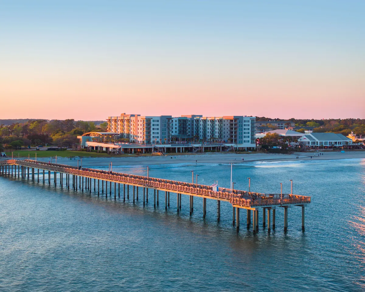 A tranquil exterior image showcasing a scenic pier stretching into the water, with colorful waterfront buildings and a beautiful sunset sky. The scene is serene yet inviting, suggesting opportunities for walks, fishing, and photography at the water’s edge.