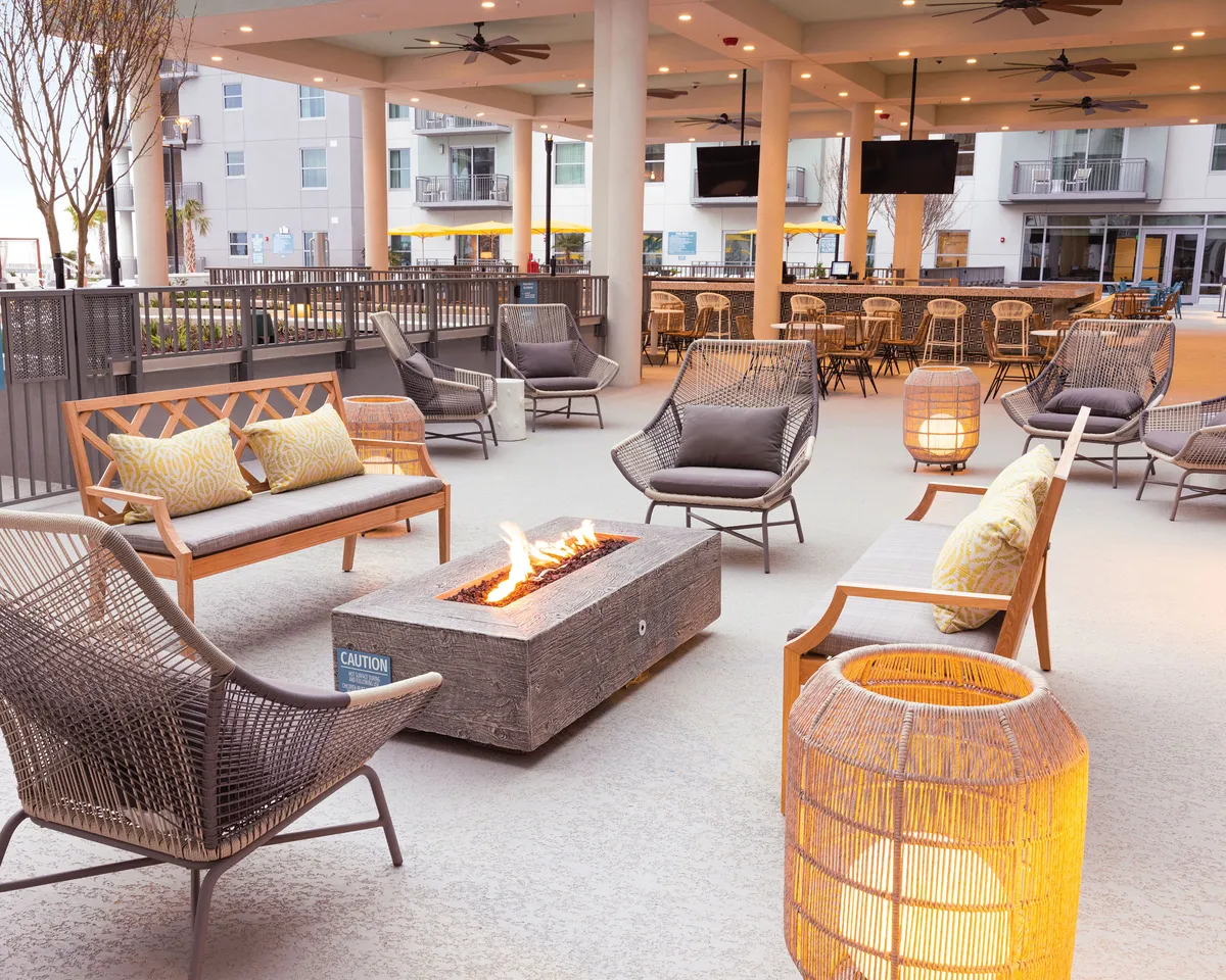 An inviting exterior fire pit area is surrounded by comfortable seating and decorative lanterns, creating a cozy and communal setting. The scene is designed for relaxed social gatherings, outdoor dining, or evening conversations, enhanced by the glow of the fire and atmospheric lighting.