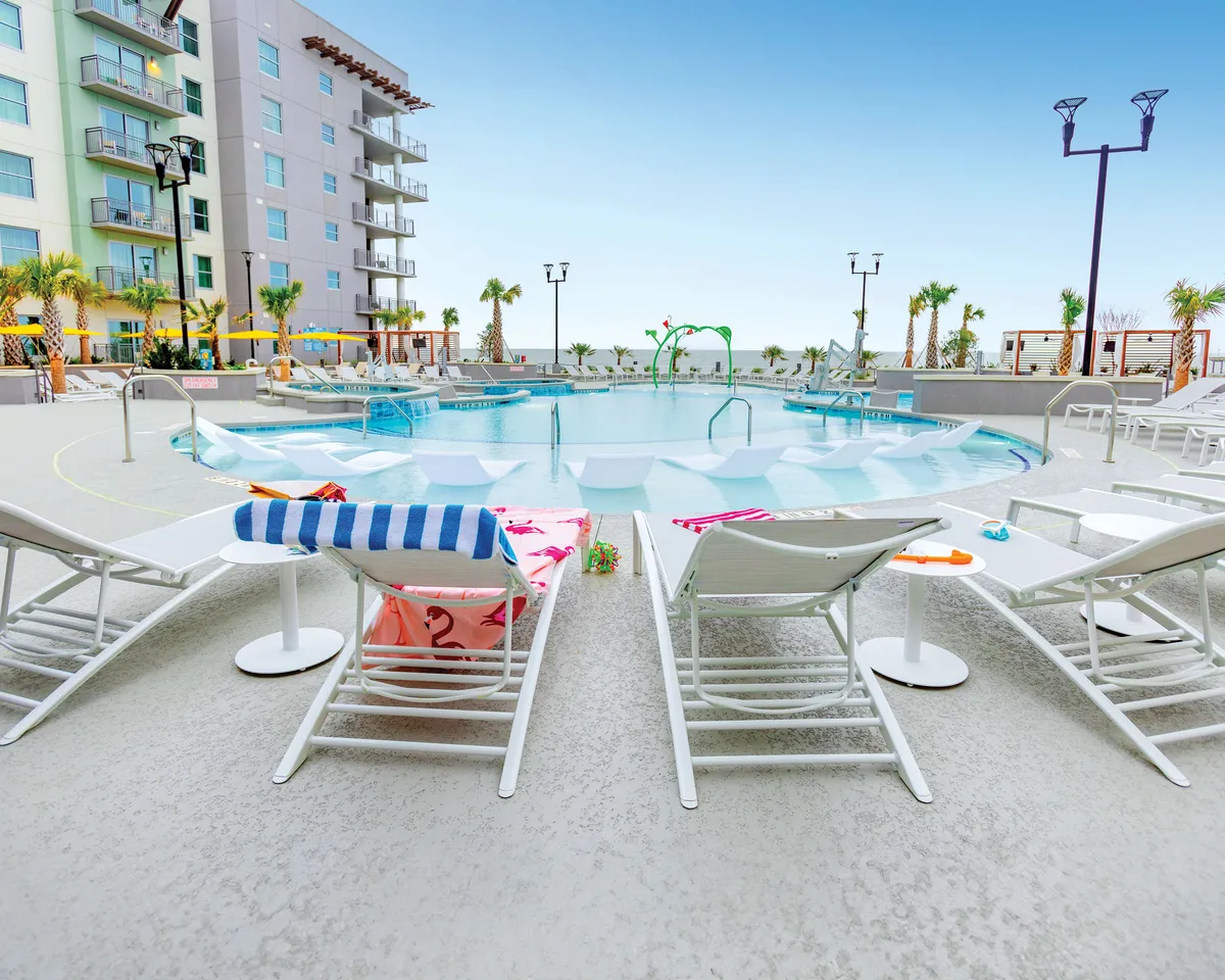 A vibrant outdoor swimming pool area is featured, surrounded by lounge chairs adorned with colorful towels and tall palm trees framing the scene. The pool is nestled next to the building facade, suggesting easy access for guests. Groups of friends are seen relaxing, sunbathing, and socializing by the pool, while sunset light casts a warm, welcoming glow.