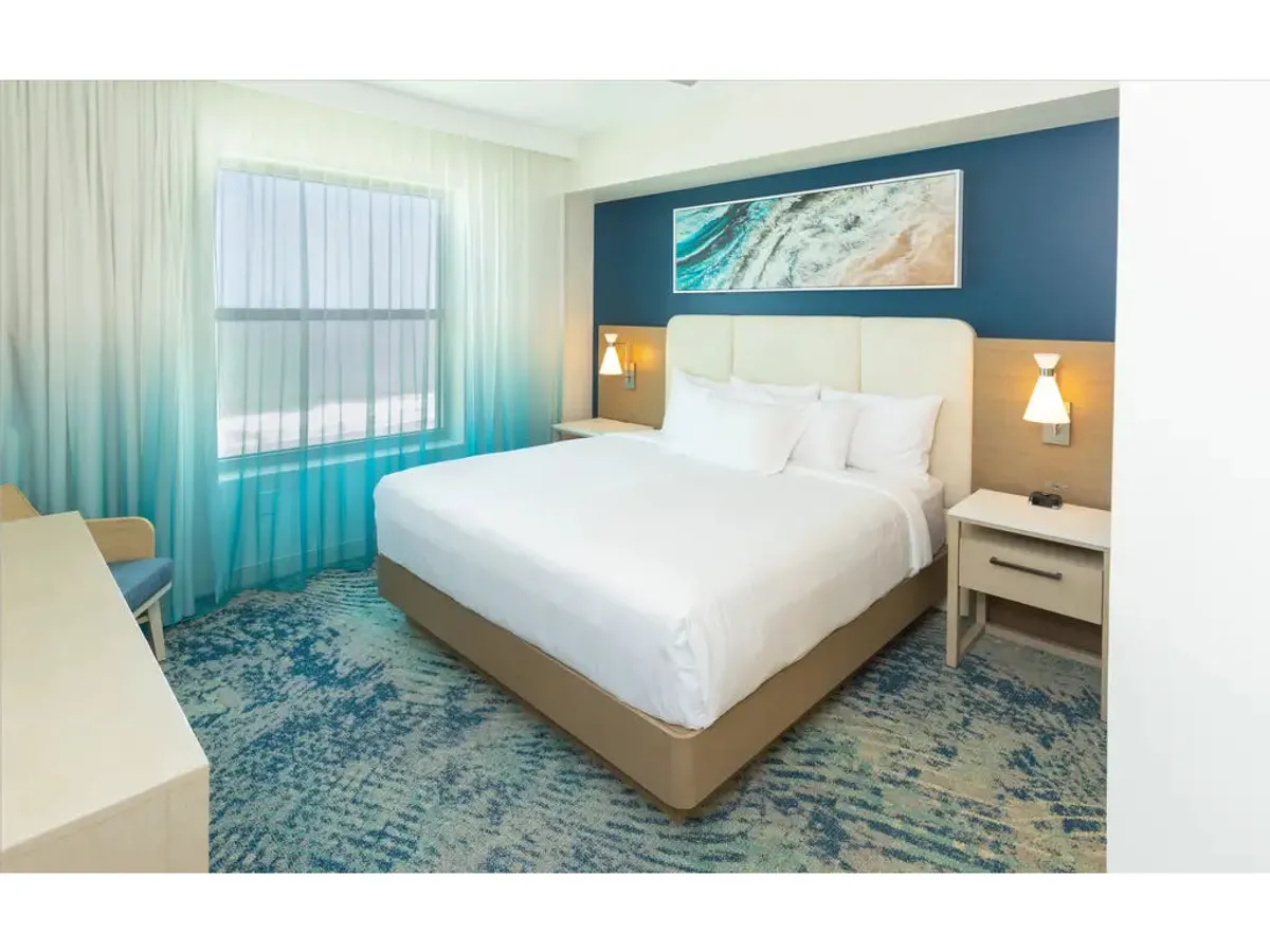 The image displays a serene king bedroom featuring a plush king bed dressed in luxurious linens. Colorful artwork adorns the wall, while soft curtains frame large windows that allow natural light to gently illuminate the space. The overall ambiance is tranquil, inviting relaxation and quiet moments.