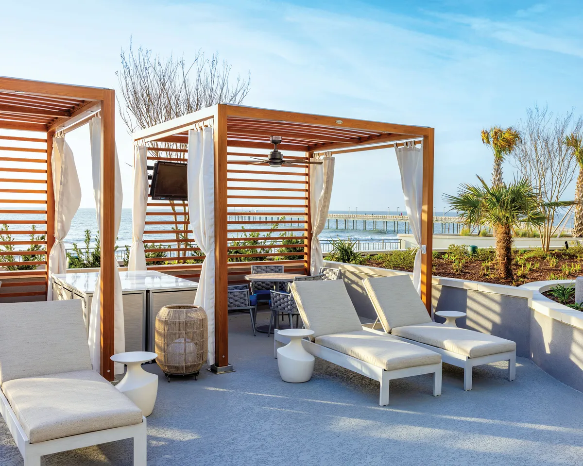 A row of stylish wooden cabanas lines the edge of the pool, each outfitted with plush lounge chairs and overhead fans. The scene overlooks the waterfront, creating a breezy, relaxed atmosphere perfect for private lounging or group gatherings. Sunset light bathes the space, emphasizing the comfort and exclusivity of the cabana experience.
