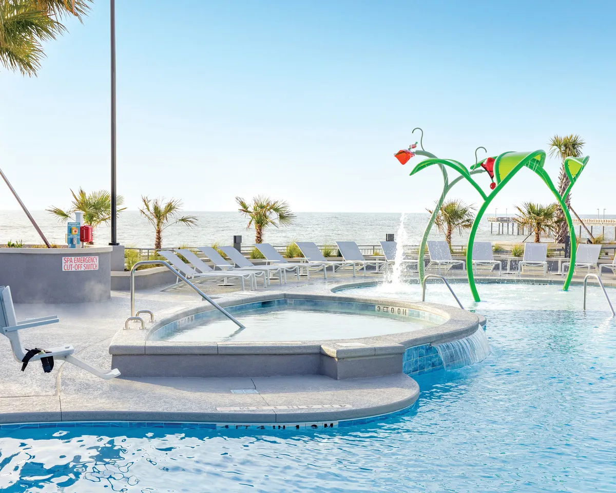 This image showcases an expansive swimming pool framed by inviting lounge chairs and features a vibrant, colorful water play structure perfect for children. The pool area is set against a stunning ocean view backdrop, with families visibly enjoying both the water and sunny weather. The scene captures playtime, relaxation, and social gatherings all in one.