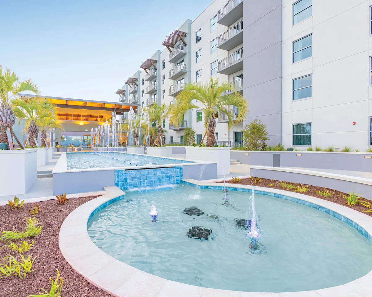 A tranquil exterior scene featuring a sparkling pool surrounded by palm trees and modern resort architecture. The area is bathed in soft sunset light, with guests lounging poolside and ambient lighting creating a romantic, relaxing atmosphere ideal for couples.