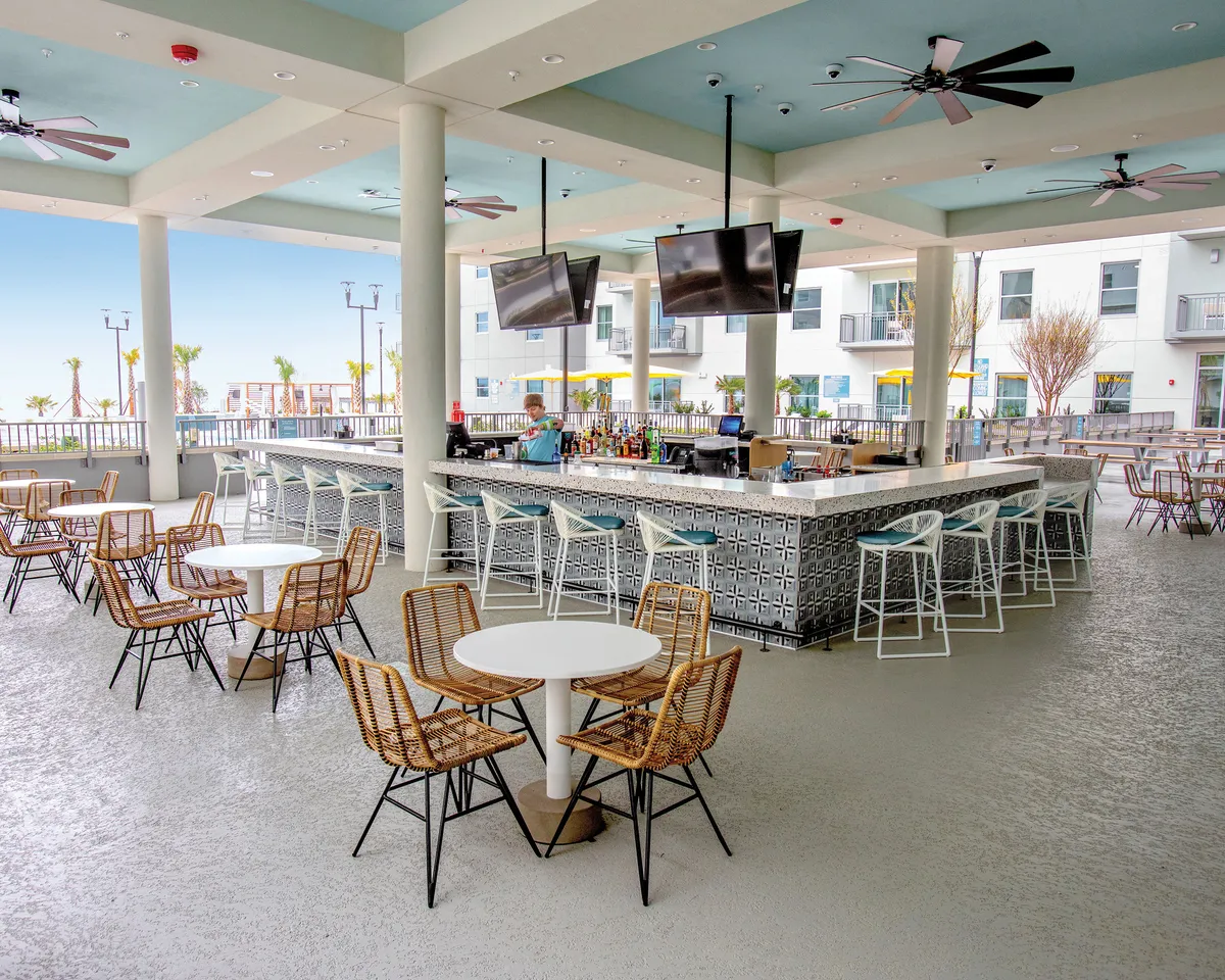 A lively outdoor bar area features a welcoming setup of tables and chairs, ceiling fans, and an open view of the resort surroundings. Guests mingle while enjoying drinks and snacks, energized by a cheerful atmosphere and the promise of sunset views from the bar.