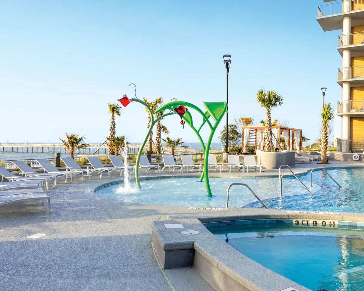 A vibrant, sunlit exterior scene featuring a colorful water play structure surrounded by a swimming pool and lounging chairs. Palm trees sway in the background, and the ocean is visible beyond the pool area. Families and children are seen enjoying the water play features, creating a joyful, energetic atmosphere.